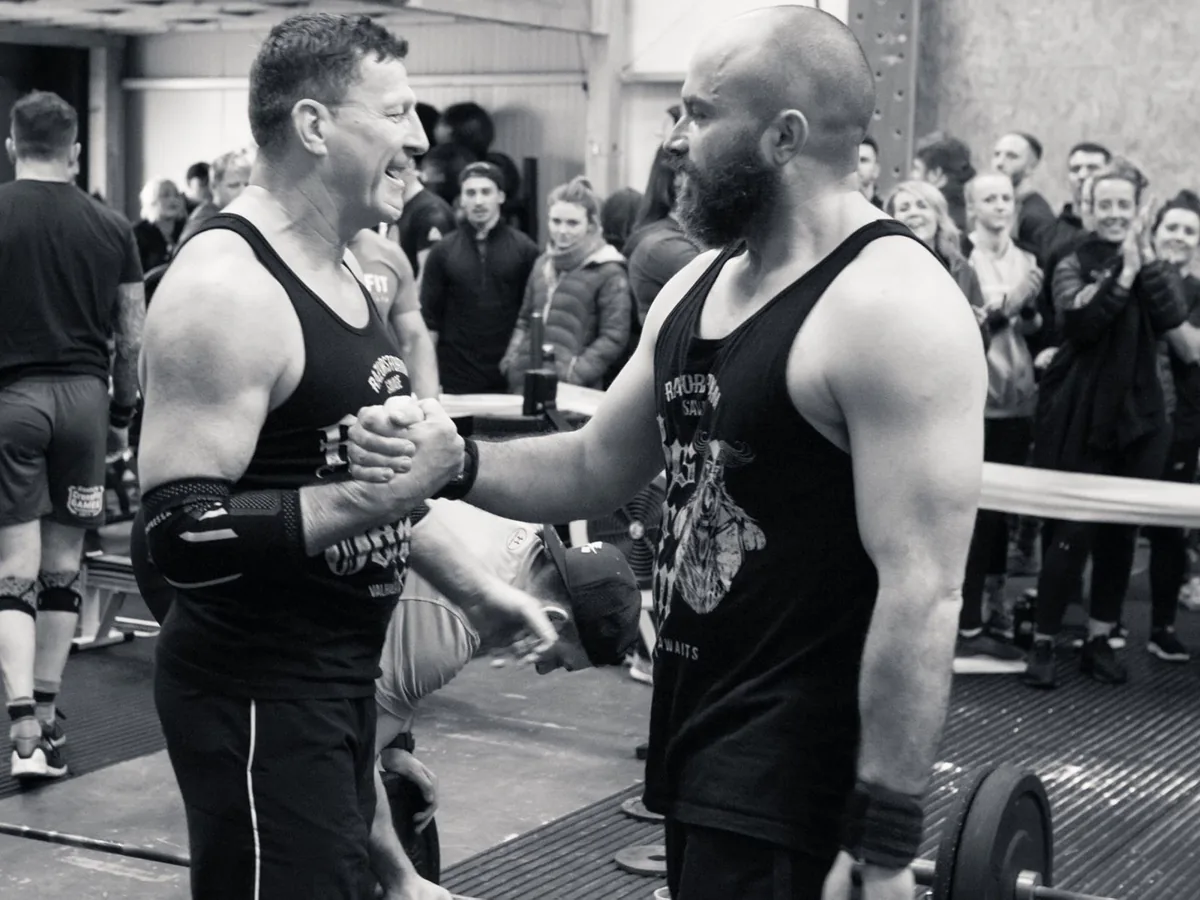 Two members shaking hands beside a loaded barbell in front of a crowd